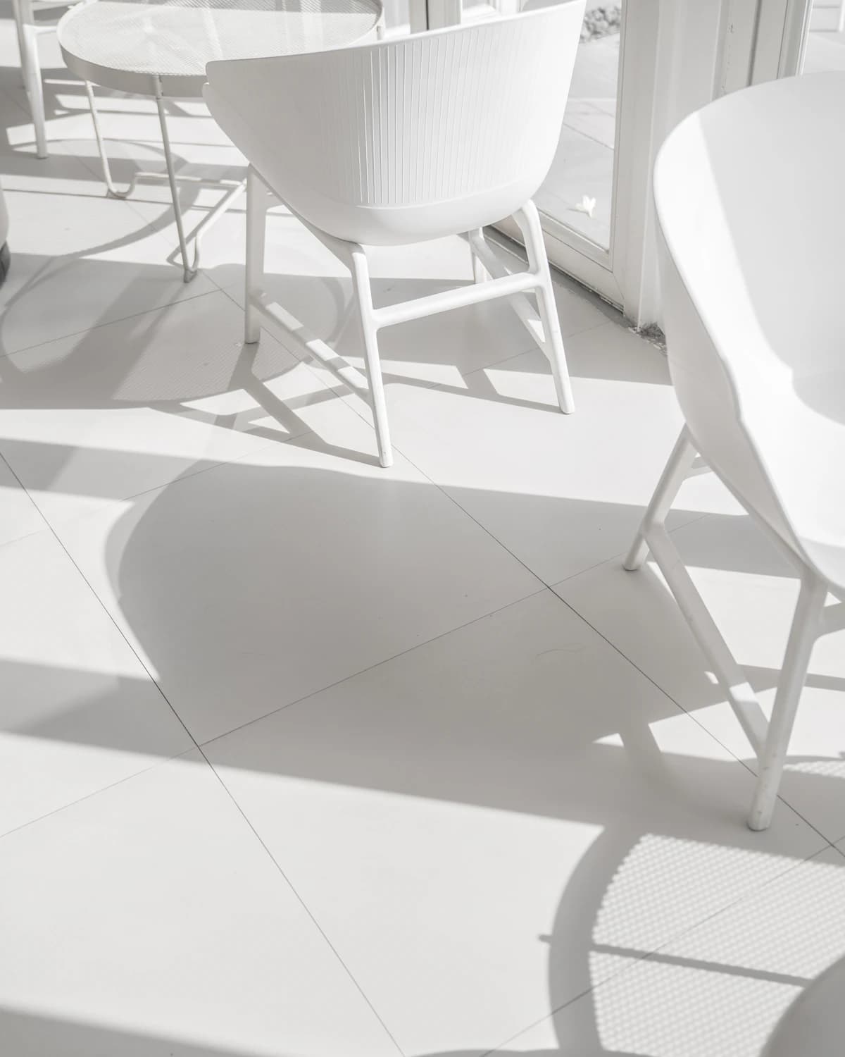 White chairs on white floor tiles flooded with midday sun, their shadows drawn sharp across the room.