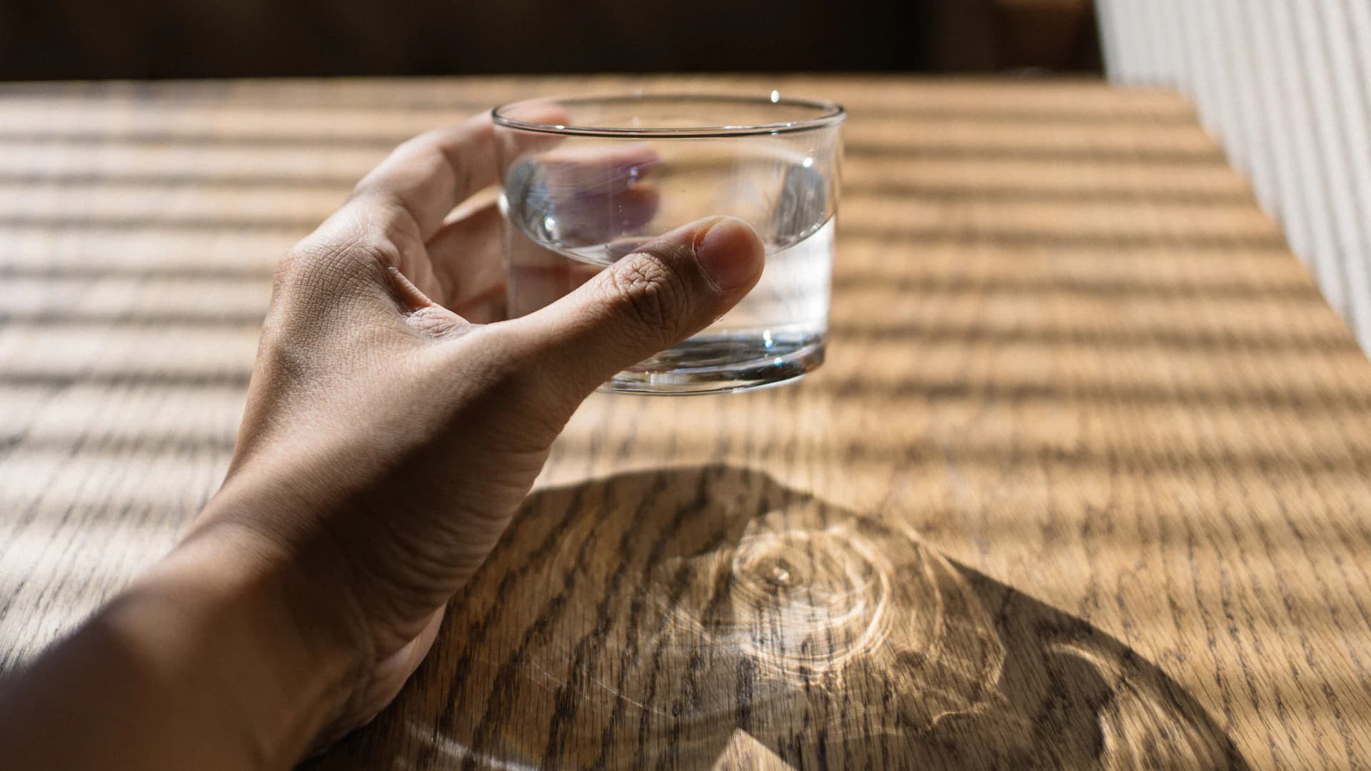 A hand holding a glass of water over a pale oak table, afternoon light through blinds casting long slatted shadows.