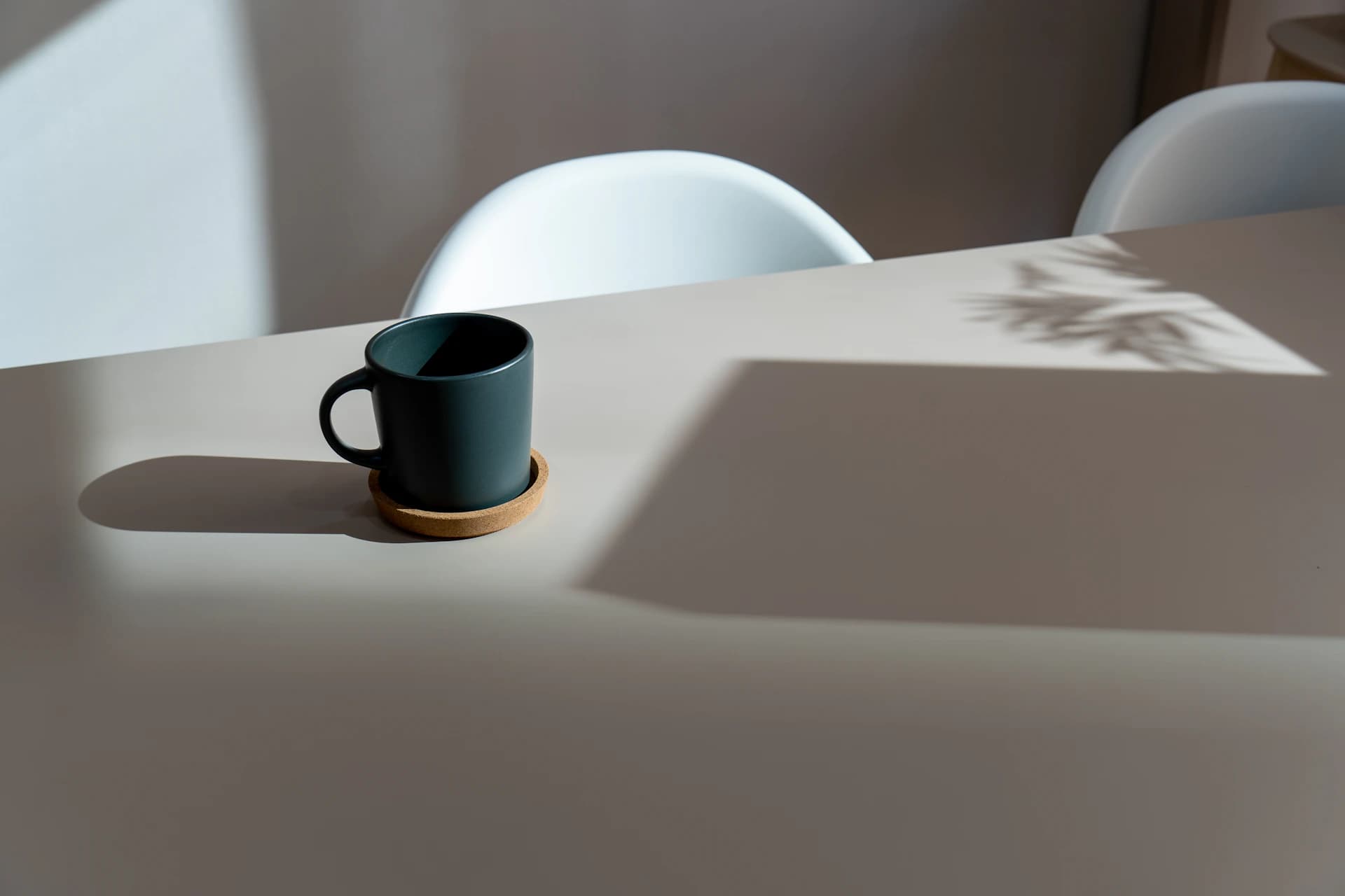 A deep-blue ceramic cup on a white table in hard morning window light, a plant throwing a crisp shadow across the surface.